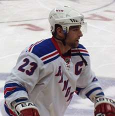 An ice hockey player shown from the waist up on the ice. He is wearing a white jersey with the word "Rangers" in blue across the front.