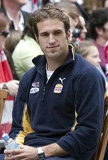 A male athlete with dark hair wearing a jumper sits in a chair.