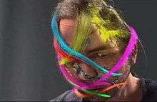 A digitally rendered version of Chris Landreth's face, slightly tilted downward, on a dark grey background. Ribbons of various colours wrap around his head in various orientations.