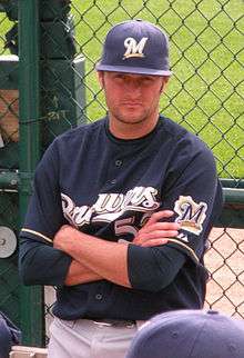 A man wearing gray pants, a navy blue baseball jersey with "Brewers" written across the chest in white and gold letters, and a navy blue cap stands with his arms crossed