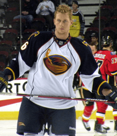 An ice hockey player stands directly upright holding an ice hockey stick horizontally across his stomach. He is wearing a no helmet and is wearing a black and white uniform with a large orange bird with an ice hockey stick on his chest.