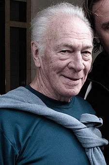 Upper torso of a gray-haired old man. He is wearing a teal T-shirt with a grey sweater tied around his neck