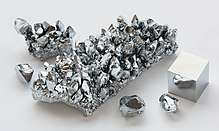 A silvery finger of chromium irregularly encrusted with diamond-like chunks of chromium of varying size. There is also a one-third sized version of the finger and three roughly hewn gem-like chunks of chromium, as well as the cube. There is a partial reflection of one of the three gem-like chunks in one of the faces of the cube.