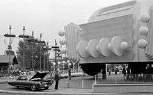 Car with its hood up in front of a World's Fair pavilion