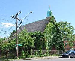 An ivy-covered building with a pointed roof and a small green dome with a cross seen from across a street, with a car parked in front. There is a chainlink fence around it and the ground slopes downwards towards its rear. A street sign at the corner reads "Colonie" and "North Pearl".
