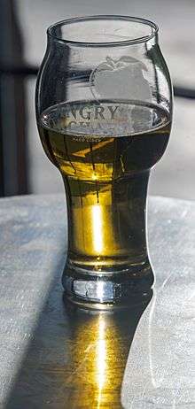 A drinking glass with the Angry Orchard name and logo on it, bulbous at the top, half-filled with clear dark yellow liquid through which the sun shines, reflected on the mental breakdown in front of it