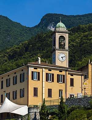 Two yellow two-story buildings, one with a clock tower reading 5:12, seen from a low angle. Behind them is a forested mountain and blue sky.