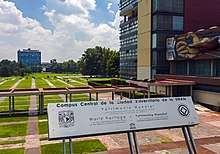 A silvery metal sign on supports leaning away from the camera saying "World Heritage Site" in Spanish and English, with supporting text. Behind it is a building with a mural, a covered walkway, and a large quad with buildings in the distance