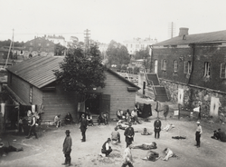 A vantage-point picture of a prison camp at the Suomenlinna Fortress in Helsinki. Around 25 Red prisoners are present in the courtyard, surrounded by a shack and a garrison building.