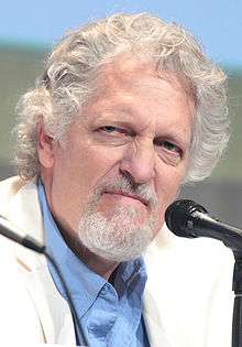 A white man with a short white beard, and white and grey curly hair. He wears a white suit with a blue shirt. The man stares away from the camera looking angry.