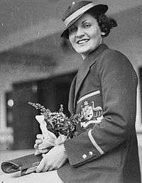 Young woman with short wavy hair, in a straw circular hat with ribbon, wearing a ceremonial blazer with the Australian coat of arms, and holding a bouquet of flowers and paper documents.