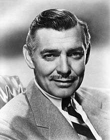Black and white promo photo of Clark Gable in 1940—a middle-aged white man with mustache and straight gray hair combed to the side, wearing a suit and smiling.