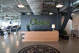 The front desk at the Classy offices: an image of an open concept office with a reception desk in the foreground. The Classy company logo is made out of plants on a wall behind the desk. The office is a modern office building designed to look like a converted loft, with large structural beams, brick walls and the HVAC pipes exposed. Many people sit at computer desks along a bank of windows in the background.