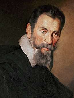 Head of a heavily bearded short-haired man with a serious expression, leaning slightly forward and facing semi-right, although his eyes look straight ahead. A white collar over a dark coat or cloak is also visible.