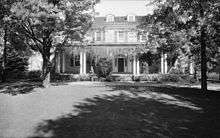Black and white image of a two story house with dormer windows on the roof and a porch with vines on the ground floor. A standing man in a suit and two large trees are in front of the house.