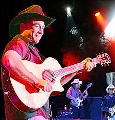 A man wearing a brown cowboy hat and red shirt, playing a guitar