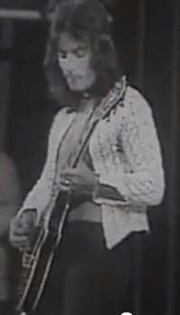 Three-quarter body shot of a 24-year-old man who is playing an electric guitar. He wears over shoulder length hair, a white shirt with chest bared and dark pants. His left hand is down the fret board, his right hand holds a plectrum and strums the strings.