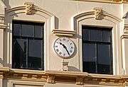Close-up photograph of a wall-mounted clock with a white face and black Roman numerals, on a pastel-yellow wall.