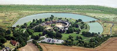 Aerial view of the fort showing the fort itself at the centre, surrounded on three sides by a moat. An area of marshy grassland bordered by the river is visible in front of the fort. Behind it are trees, fields, a car park and a church.