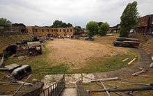 Wide view of the fort's interior, showing a semi-circular sandy area in which items of military equipment are standing. A row of brick buildings is visible at the rear.