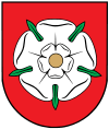 A coat of arms depicting a white flower in the middle that has a yellow stamen and green leaves all on a red background