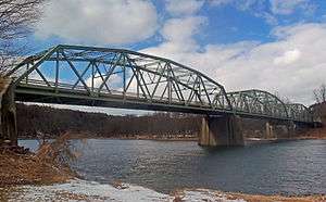 A three-span bridge with concrete piers at the joins and green-painted metal overhead arching latticework structures is seen from the shore of the river it crosses, below its deck. There is some snow on the ground but the river is not frozen. Both sides are wooded.