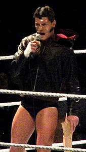 A dark-haired Caucasian male stands in a wrestling ring with grey ring ropes. He is wearing short black wrestling tights with a black, hooded jacket, and his face is covered with a clear plastic mask. He is holding a microphone to his mouth with his right hand, and in his left hand he is holding brown paper bags.