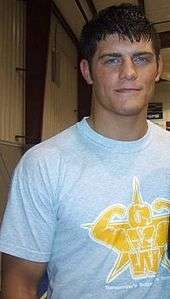 A close-up of a young Caucasian male with black hair. He is wearing a light-colored tee-shirt with the yellow logo of the Ohio Valley Wrestling promotion upon it.