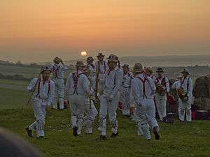 A group of twelve individuals wearing all-white costumes, including hats, are dancing in the centre of a green, grassy space. The low position of the sun indicates that it is early in the morning