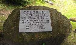 A circular boulder takes up most of the image; behind it can be seen green grass. On the boulder is a light-grey plaque. On this plaque are written the words: "Coldrum Stone Circle has been vested in The National Trust as a Memorial to Benjamin Harrison 1837–1921 of Ightham 10 July 1926". All of the letters are in capital letters; those stating "Coldrum Stone Circle" are larger than the others.