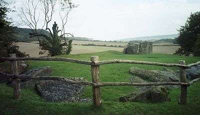 A rural scene. Immediately in front of the image is a timber fence, behind which lie several large grey stone boulders in a field.