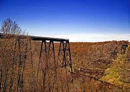 A trestle bridge across an autumnal valley at left ends in a drop off at center, with collapsed remnants at right, all under a bright blue sky