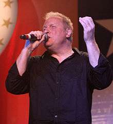A blond-haired man wearing a black shirt, singing into a microphone