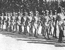 Soldiers in colonial-era military uniform march with rifles shouldered.