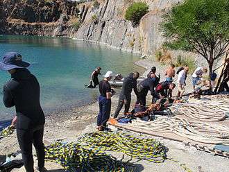 A group of about 12 divers on the shore of a flooded quarry preparing surface-supplied diving equipment for diver training exercises. Several umbilicals are laid out for use in figure 8 coils.
