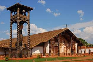 A wooden bell-tower and a church in three-quarter view. The bell-tower consists of a roof-covered platform supported by four columns with twisted fluting. Clocks are attached to the platform and a spiral staircase leads to it. The facade of the church is white and decorated with orange paintings. The church roof is rather large.
