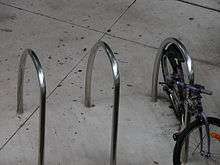 Three stainless steel U-rings set in concrete, without bolts. A bicycle is chained to the one on the right.