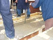 A handheld concrete vibrator consolidates fresh concrete in a wooden form for a concrete beam.