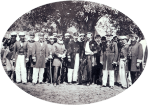 A photograph showing a large group men, most of whom wear uniforms, standing beneath shade trees