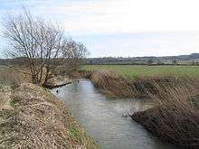a Lowland landscape in the wide Welland valley, with distant hills on the horizon.  Most of the picture is taken up with what appears to be a river flowing from in front of the viewer and out through the top of the frame, with a smaller stream joining from the right.  But by convention it is the Welland that flows in from the right and out the top, and the Eye Brook that is at the photographer's feet and loses its identity at the junction with the Welland.  It is a winters day, the trees on the left bank are bare and the grass brown along the waters edge.  But in the middle distance a field of winter wheat is verdant.