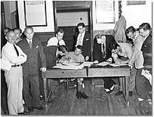 A soldier is sitting at a wooden table, with two men signing papers at the table and a line of men lined up on the right. On the left are two men, one in shirt and tie and the other in a suit facing the camera.