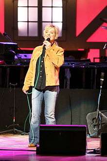 A blonde woman wearing a tan jacket, black shirt and blue pants, singing into a microphone