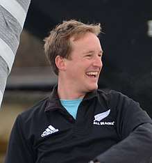 A photo of Colman, dressed causally, laughing. He stands beside a furled foresail.