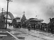 Black and white photo of a dockside scene. Buses are driving in front a crowd of people. An aircraft carrier and a bridge are visible in the background.