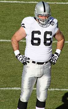Color photo of Cooper Carlisle wearing football helmet and white, silver and black uniform of Oakland Raider, standing with hands on hips.