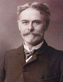 Head and shoulders of a middle-aged man who is looking at the viewer: He has a moustache and goatee, and his hair is short and parted in the middle. He is wearing a formal jacket, with a bow tie and wingless collar.