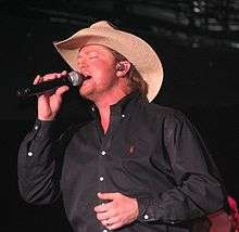 A man in a black shirt and cowboy hat, singing into a microphone