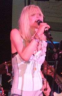 A female musician, Courtney Love, singing into a microphone at a concert. She is wearing a lingerie corset and has long blonde hair.