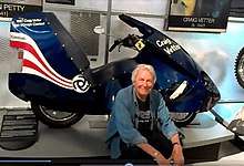 Craig Vetter, a gray haired man wearing a blue open shirt sitting just below his dark blue 1981 Streamliner motorcycle exhibited at a show in 2016