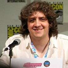 A Caucasian male wearing a white shirt sits in front of a microphone, smiling. He has large, curly brown hair which almost covers his eyes.